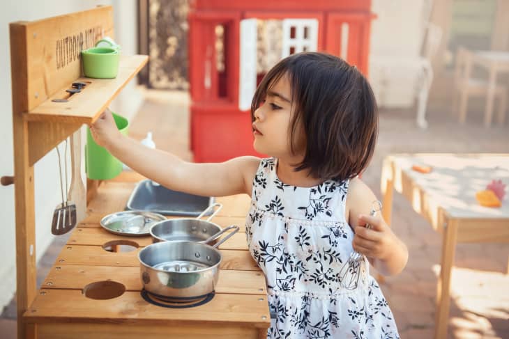 A toddler girl plays with her outdoor kitchen in her backyard on a warm, sunny day. There are pots and pans and kitchen tools and a table and chairs in the background and also a red playhouse.