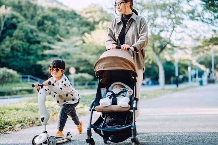 Girl with sunglasses riding a scooter with her mother pushing a baby stroller
