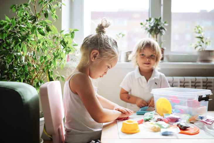 Sister and brother, sibling children molding from modeling clay together at the table. Sibling relationships and joint creativity at home in a sunny apartment.