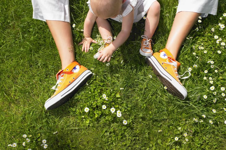 shot from above, a baby sitting inbetween someone's legs on the grass. grass has little white flowers. baby is wearing white clothes and orange hi-top sneakers. you only see the adult's legs and they are wearing orange hi-top sneakers as well.