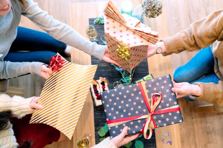 Three people exchanging wrapped gifts with festive bows, sitting around a table with snacks and drinks.