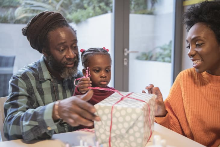 Mature man with granddaughter sitting on his lap, girl pulling ribbons on gift, mother smiling