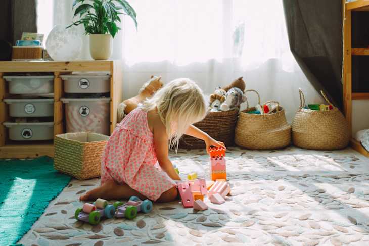 A young child in a pink dress plays on a soft rug, stacking colorful blocks beside toy baskets and a potted plant.