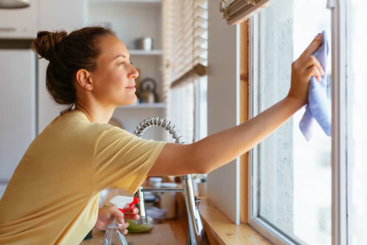 Side view of smiling young housewife with dispenser in hand washing window glass in kitchen during routine work at home