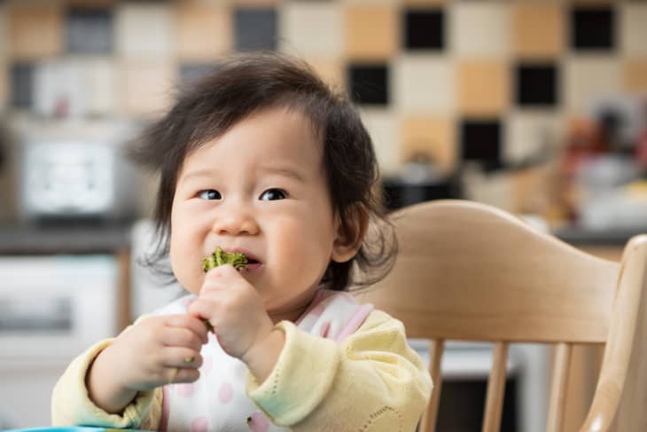 Toddler in high chair eating broccoli in a kitchen with checkered backsplash.