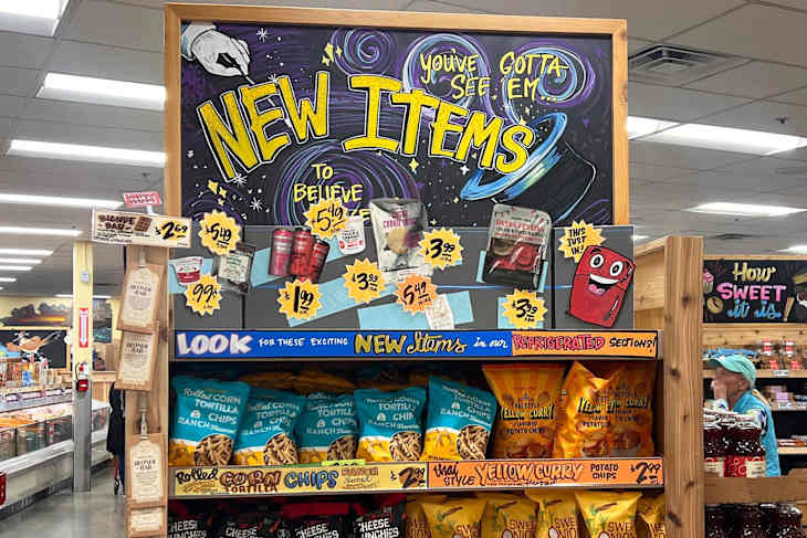 Display of new snack items in a grocery store, featuring colorful packaging, signage, and a shopper in the background.