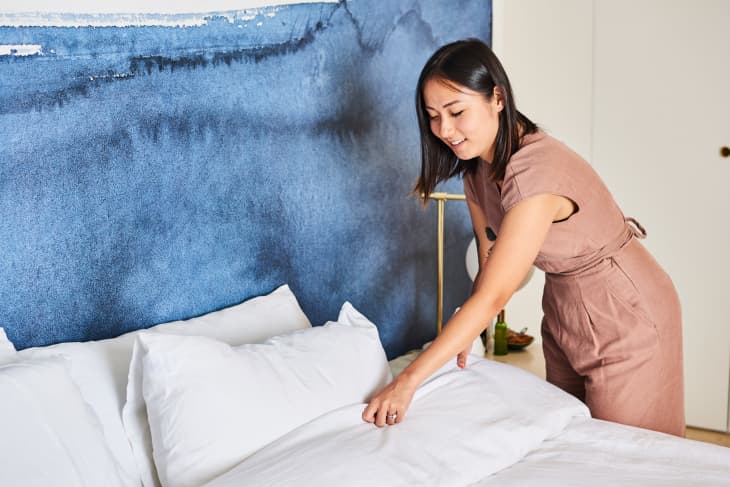 Woman in a brown dress making a bed with white sheets against a blue abstract wall.