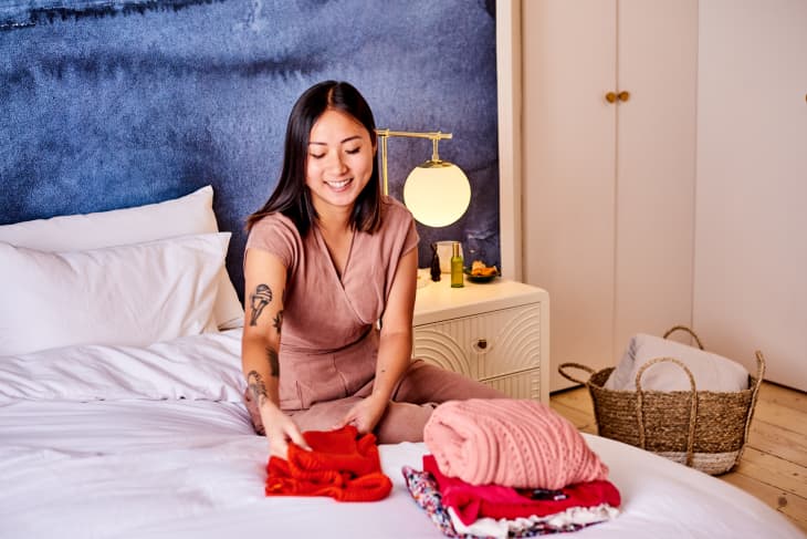 Woman folding clothes on a bed in a cozy bedroom with a blue accent wall and a bedside lamp.