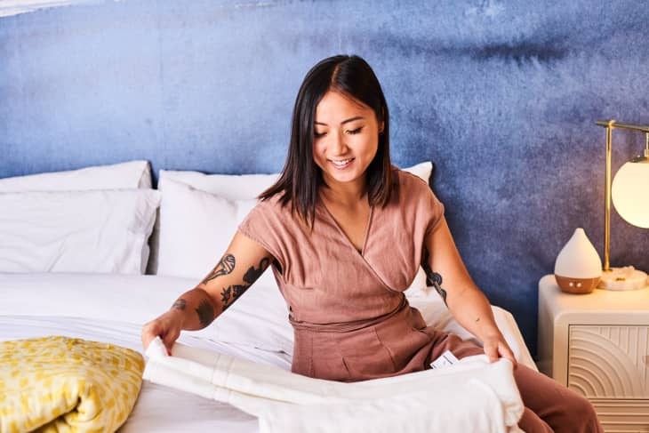 Woman in a brown dress making a bed with white sheets, blue wall background, and a bedside lamp.
