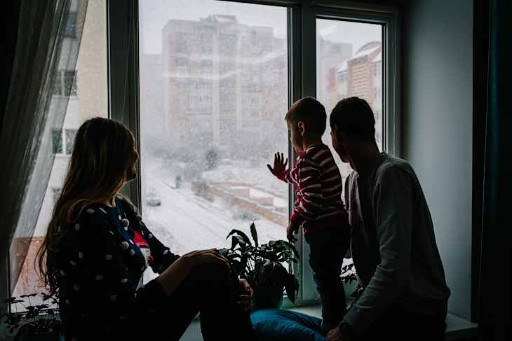 Merry Christmas and happy holidays! Happy family: mom, dad and little son in Santa Claus sweaters sitting on window and looking at the winter park. Room decorated on Christmas. Enjoy the snowfall.