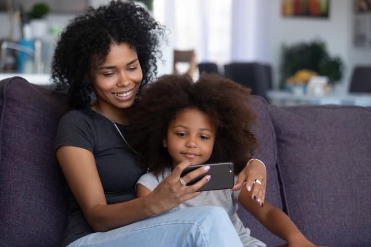 Happy smiling african american mother and little daughter taking selfie on smartphone, mixed race mom and child girl making video call on cellphone, mommy kid watch cartoons using phone app at home