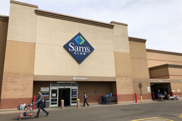 The exterior of a Sam’s Club store with a shopper walking in and away from it.