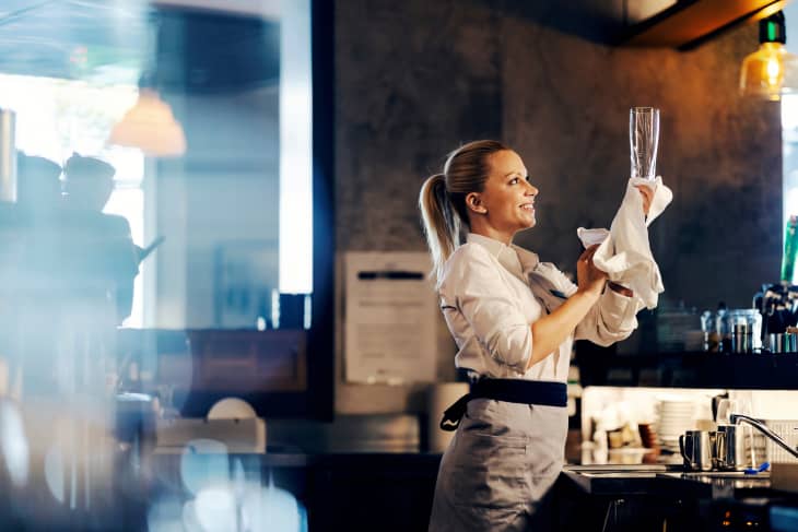 A smiling waiter cleans a glass while standing at a bar.