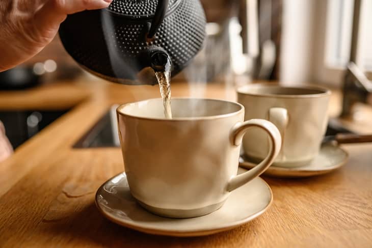 Person pouring hot water from kettle in cup at kitchen to prepare tea. Teapot and ceramic mug with warm beverage