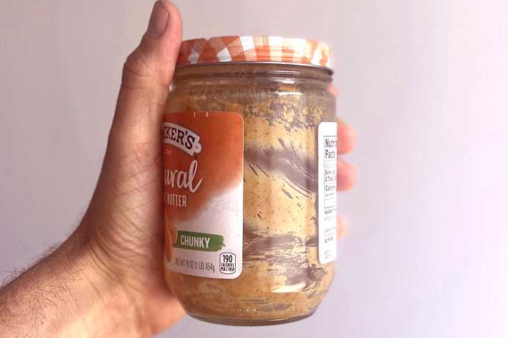 A hand holding a jar of chunky natural peanut butter with a striped lid and nutrition label.