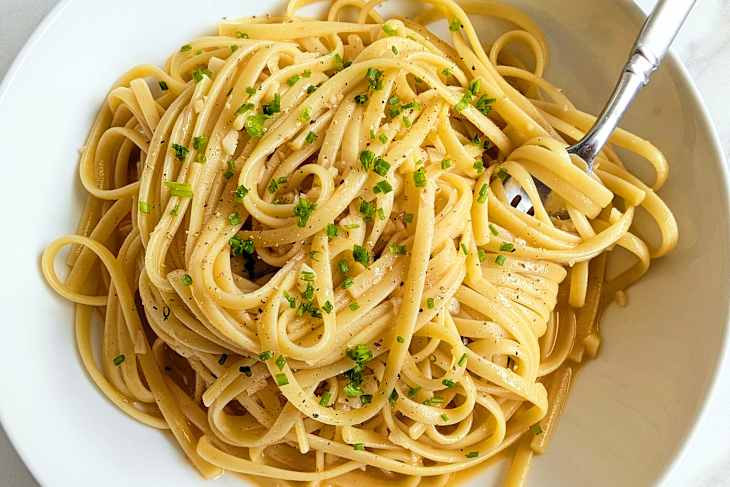 Linguine pasta topped with chopped chives and black pepper in a white bowl, with a fork resting on the side.
