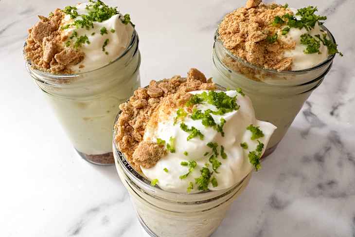 Three jars filled with creamy desserts topped with whipped cream, crushed cookies, and green zest on a marble surface.