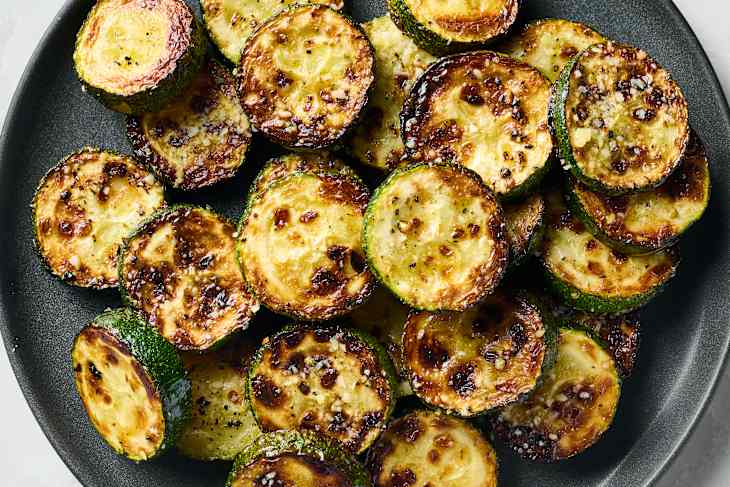 Sautéed zucchini rounds with a golden-brown crust, garnished with seasoning on a dark plate.
