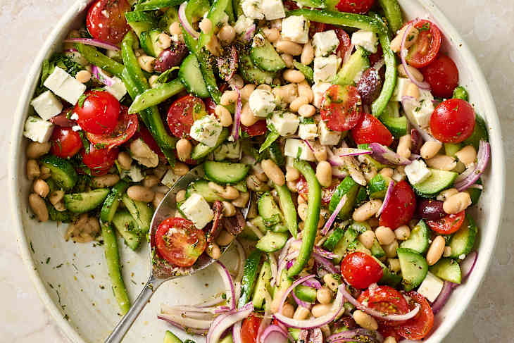 A colorful salad with cherry tomatoes, green bell peppers, cucumbers, red onion, feta cheese, and white beans, garnished with herbs.