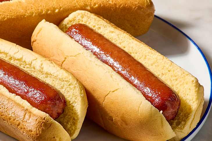 Three hot dogs in soft buns on a white plate, showcasing shiny sausage and golden bread.