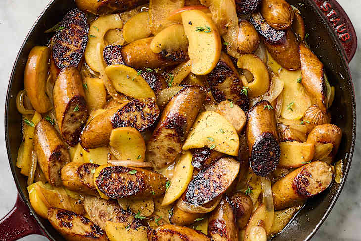 Sautéed sausages with sliced apples and onions in a cast iron skillet, garnished with fresh thyme.