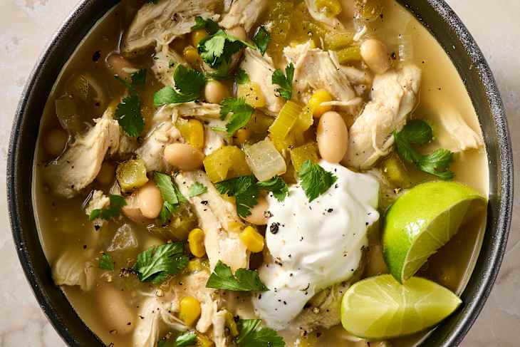 Bowl of chicken soup with shredded chicken, white beans, corn, green chilies, cilantro, and a dollop of sour cream, garnished with lime.