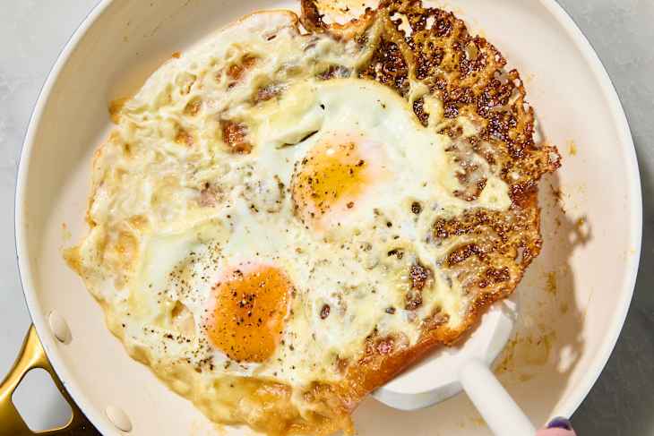 Two fried eggs in a cream-colored skillet, one with crispy edges, seasoned with black pepper.