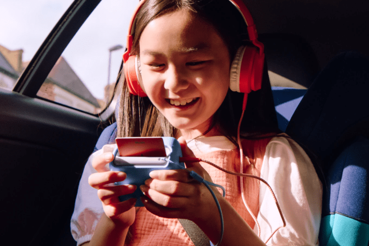 Girl in a car wearing red headphones, playing a handheld game console, smiling.