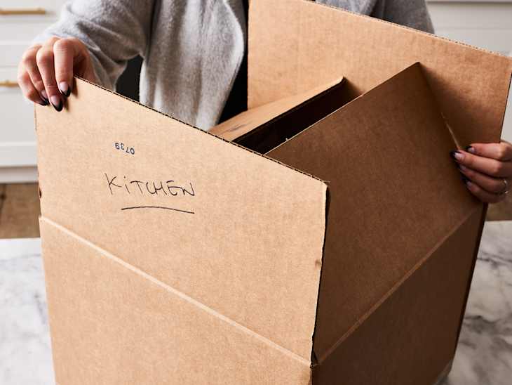 A cardboard box labeled "kitchen" that is being folded to prepare to tape for moving
