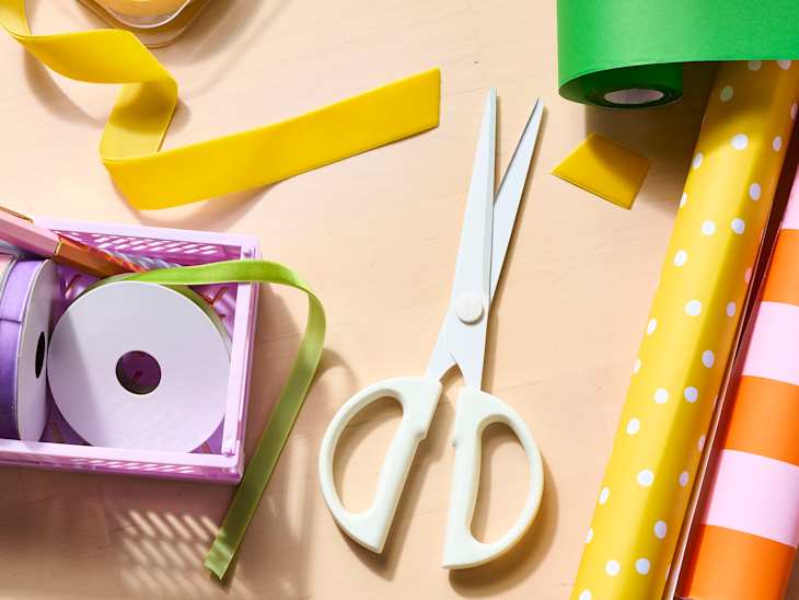 Crafting supplies on a table featuring scissors, colorful ribbons, and patterned wrapping paper rolls.