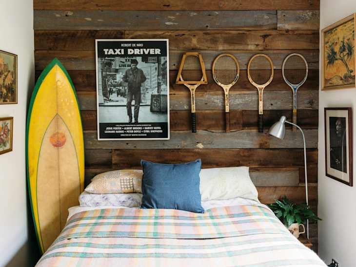 A surfboard, posters, and tennis rackets above a bed in a rustic bedroom.