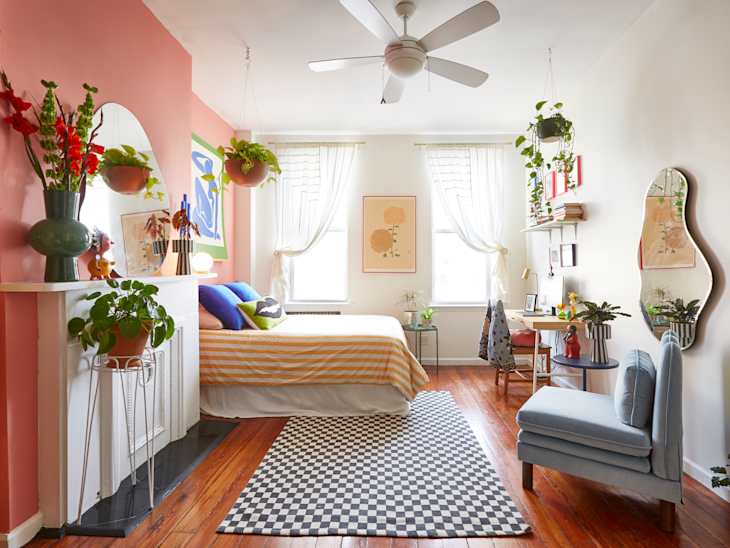 Bedroom with pink accent wall, striped bedding, plants, checkered rug, and a desk with a chair under a window.