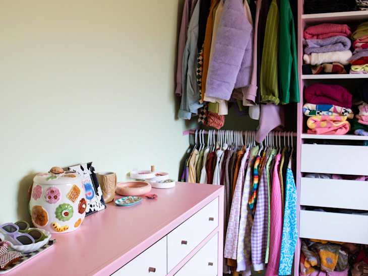 Pink dresser with decorative jars and trinkets beside a closet with colorful clothes on hangers and shelves.