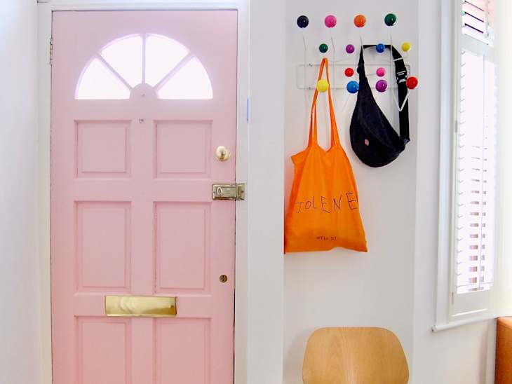 Pink front door with a window, colorful wall hooks, an orange tote bag labeled "JOLENE," and a wooden chair.