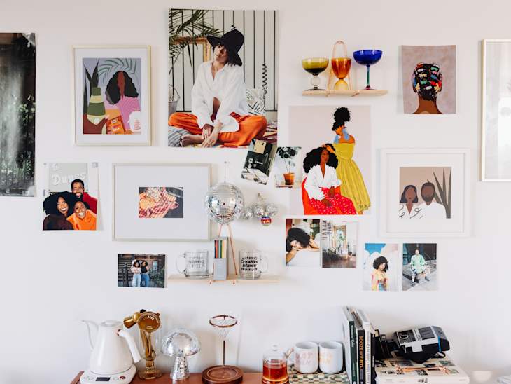 Gallery wall with colorful art prints, photos, and glassware on shelves above a table with books and a coffee maker.