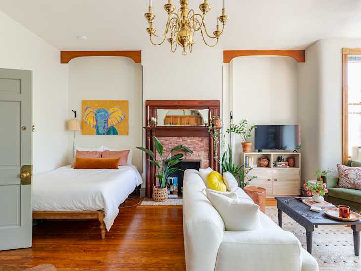 Living room with a white sofa, green armchair, TV, plants, and a bed. Chandelier and colorful artwork above the bed.