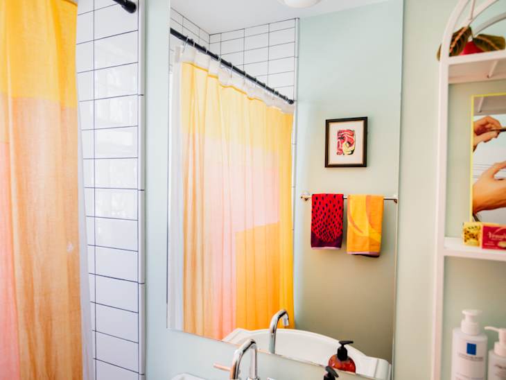 bathroom with shower with white tiles with black grout, pale orange shower curtain, pale sage green walls, shelves with grooming products, reed diffuser, white pedestal sink, red and pale orange accents