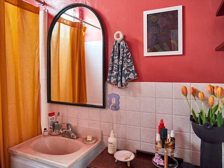 Bathroom sink area with a round mirror, yellow shower curtain, orange tulips in a black vase, and various toiletries.