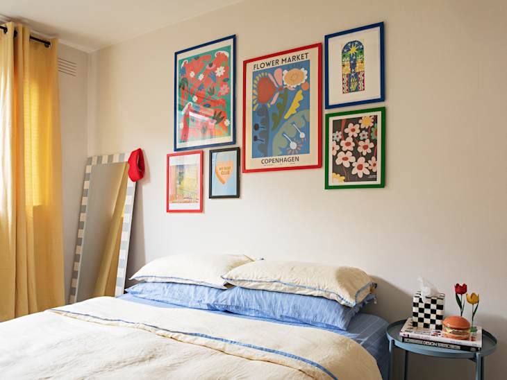 Cozy bedroom with a bed featuring blue and cream bedding, colorful wall art, a mirror, and a side table with decor.