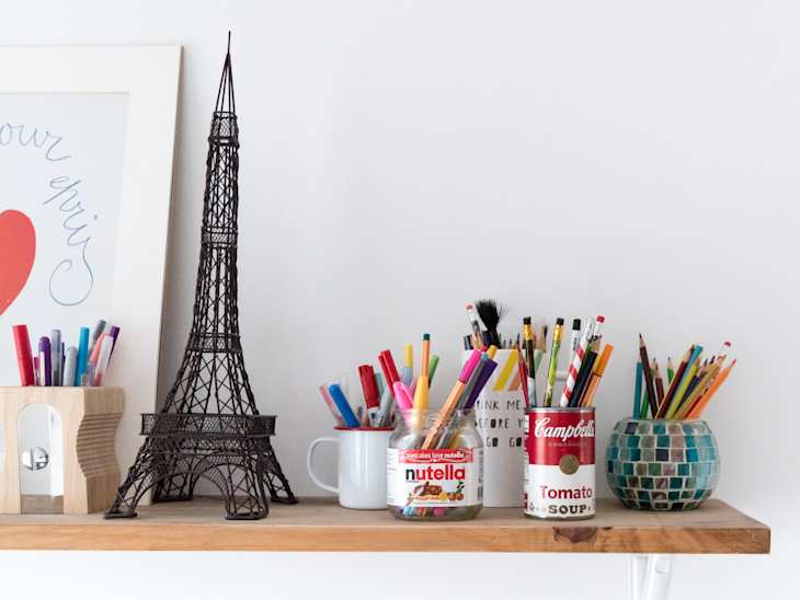 Shelf with Eiffel Tower model, framed art, and colorful pens in jars, including Nutella and Campbell's soup cans.