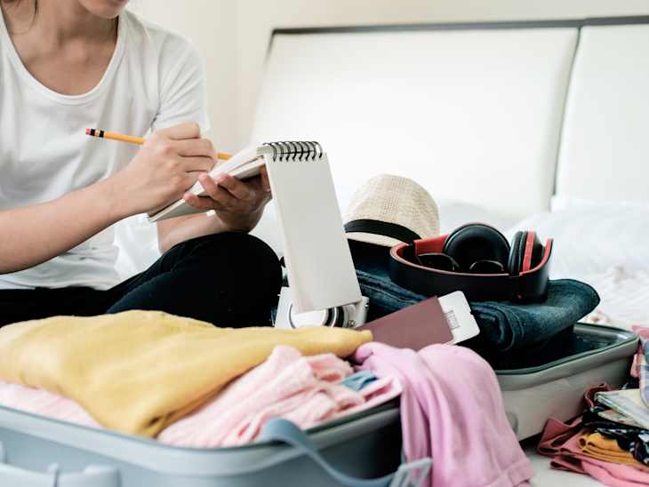 Person writing in a notebook next to an open suitcase with clothes, a hat, headphones, and a passport on a bed.