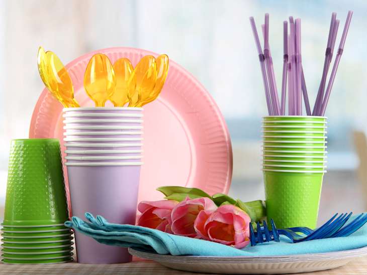 Stacked colorful plastic cups, plates, and utensils with pink tulips and blue napkins on a table.