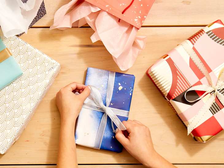 Overhead shot of two hands tying a silver ribbon onto a small package wrapped in blue and silver abstract paper.