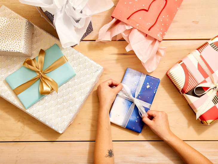 Overhead shot of someone tying a bow with silver ribbon onto a small gift in blue abstract wrapping paper, with assorted wrapped gifts on a light wood surface.