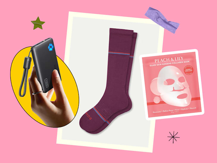 A hand holding a black portable charger, maroon socks, a pink collagen mask, and a purple hair tie on a pink background.