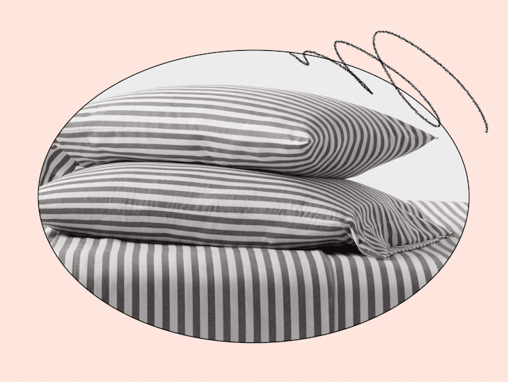 Stacked gray and white striped pillows on a matching striped bed sheet, set against a soft pink background.