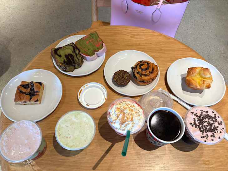 Assorted pastries and drinks on a wooden table, including green and pink swirl cakes, a chocolate muffin, and various beverages.