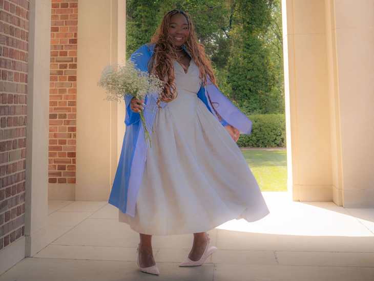 A woman in a light dress and blue graduation gown holds a bouquet of flowers, smiling in a sunlit outdoor setting.