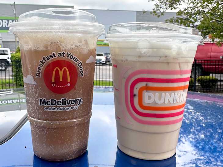 Two iced coffee drinks from McDonald's and Dunkin' on a blue surface, with a blurred background of greenery and vehicles.