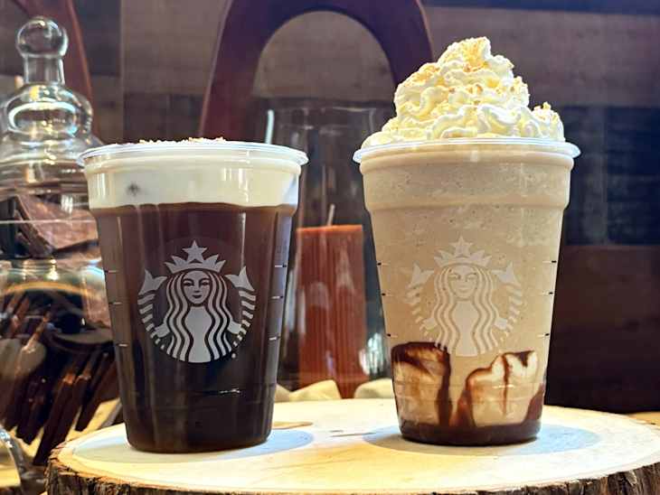 Two Starbucks beverages on a wooden surface: a dark iced coffee topped with cream and a blended drink with whipped cream and chocolate drizzle.